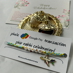 A handmade yellow and blue braided bracelet with white letter beads spelling 'RAKHI' and a small airplane charm, presented on a branded white card. Behind it is a gold-toned decorative tray with a small bottle and a floral 'Happy Rakhi' card on a light marble surface.