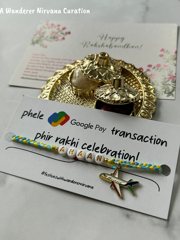 A handmade yellow and blue braided bracelet with white letter beads spelling 'RAKHI' and a small airplane charm, presented on a branded white card. Behind it is a gold-toned decorative tray with a small bottle and a floral 'Happy Rakhi' card on a light marble surface.