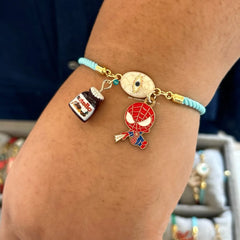 A close-up of a person's wrist wearing a light blue cord charm bracelet with gold-tone hardware and three charms: a small nutella jar charm, a round evil eye charm, and a Spider-Man character character
