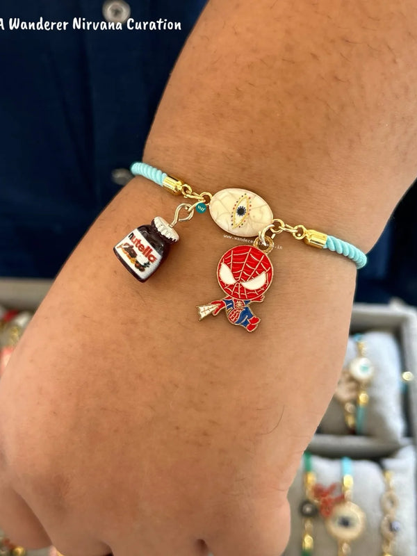 A close-up of a person's wrist wearing a light blue cord charm bracelet with gold-tone hardware and three charms: a small nutella jar charm, a round evil eye charm, and a Spider-Man character character